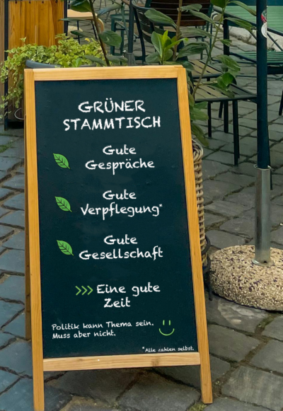 Aufsteller vor einem Café mit der Aufschrift „Grüner Stammtisch“ sowie den Punkten gute Gespräche, gute Verpflegung und gute Gesellschaft.