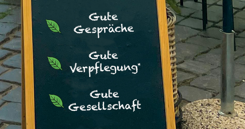 Einladung-Grüner-Stammtisch Aufsteller vor einem Café mit der Aufschrift „Grüner Stammtisch“ sowie den Punkten gute Gespräche, gute Verpflegung und gute Gesellschaft.